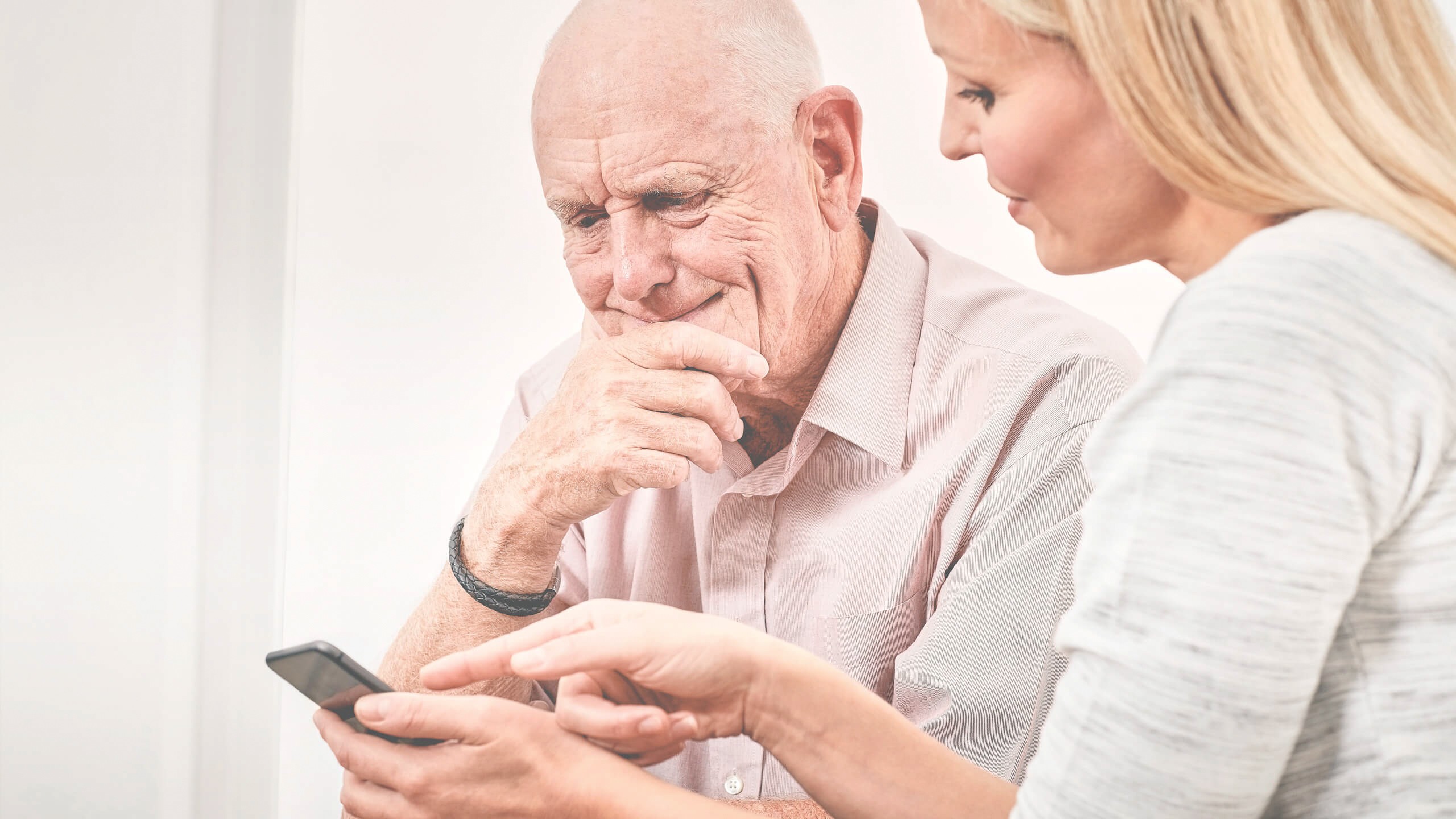 Eine ältere Person schaut nachdenklich auf ein Smartphone, während eine jüngere Person neben ihr sitzt und ihr etwas auf dem Gerät zeigt. Die Szene vermittelt den Eindruck, dass die jüngere Person der älteren Person hilft, das Smartphone zu verstehen
