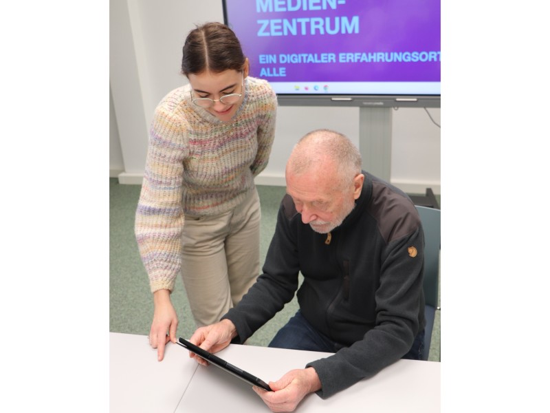 Mitarbeiterin erklärt älterem Mann etwas am Tablet.