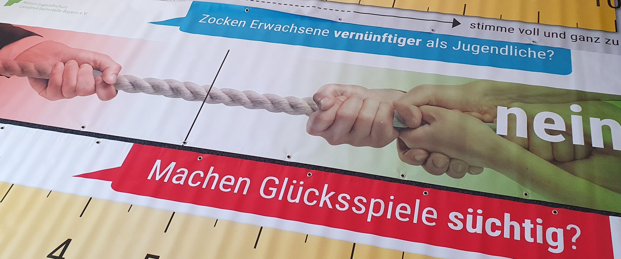 Plakat mit Skalen und Fragen zu Glücksspielsucht, begleitet von einem Bild zweier Hände im Tauziehen.