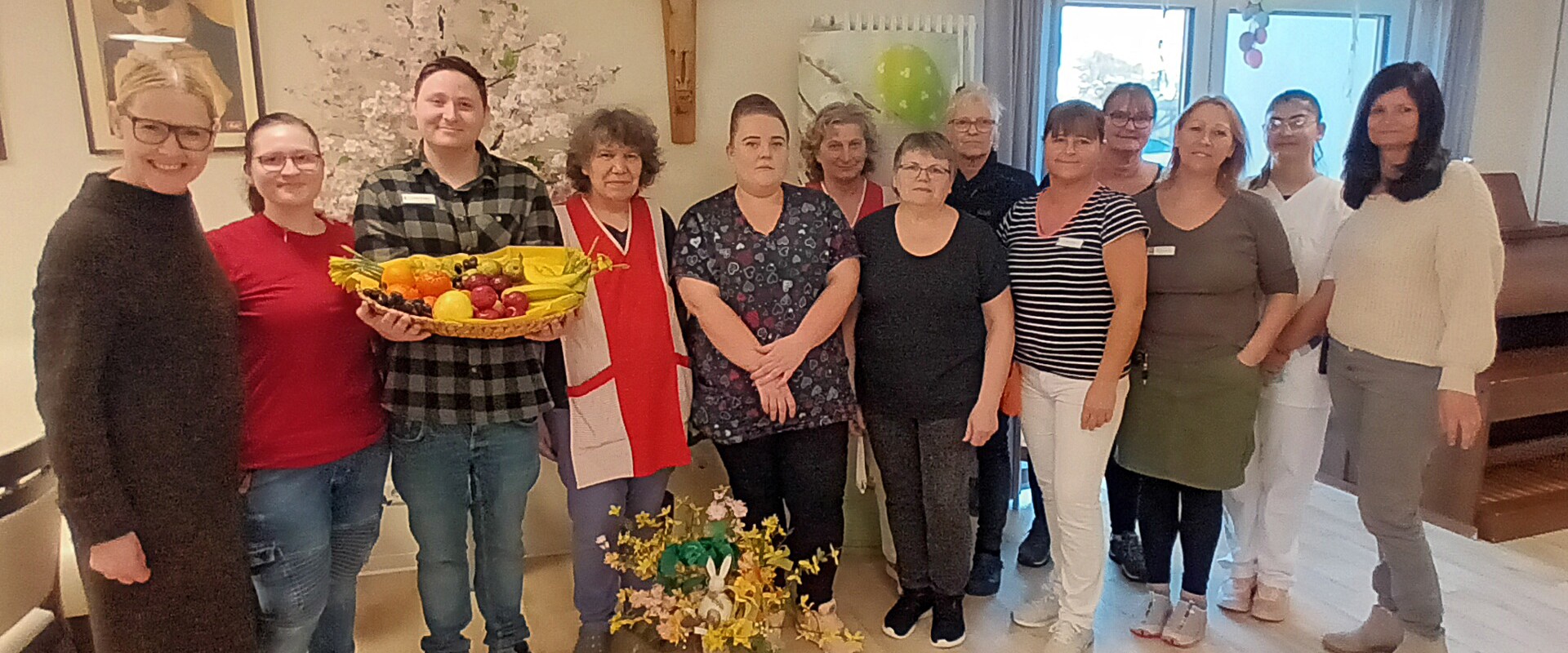 Eine Gruppe von zwölf Personen posiert lächelnd, eine Person hält einen Korb mit frischem Obst in der Hand.