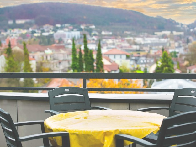 Rundtisch mit Stühlen auf einer Terrasse, die auf eine hügelige Stadtlandschaft blickt.