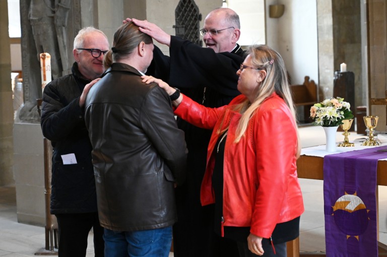 Ein Pfarrer segnet einen Mann in einer Kirche, während zwei weitere Personen ihm die Hände auflegen.
