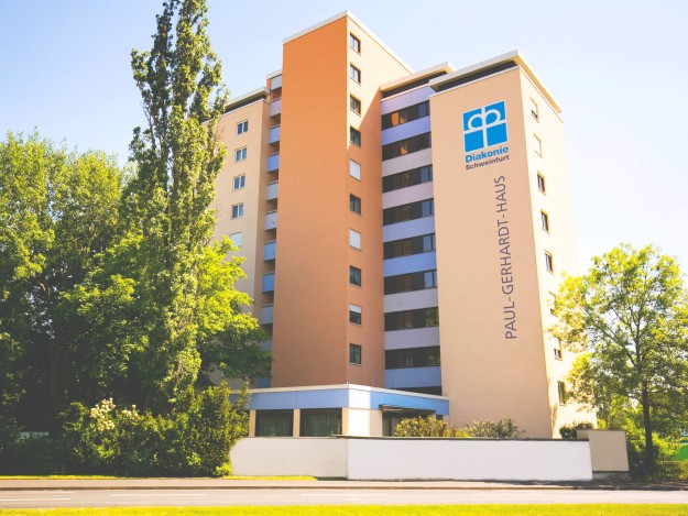 Ein mehrstöckiges, hellgelbes Gebäude des Paul-Gerhardt-Hauses, flankiert von grünen Bäumen und der Beschriftung „Diakonie Schweinfurt“ auf der Fassade.