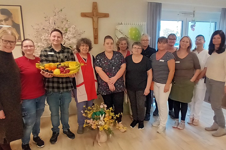 Eine Gruppe von zwölf Personen posiert lächelnd, eine Person hält einen Korb mit frischem Obst in der Hand.