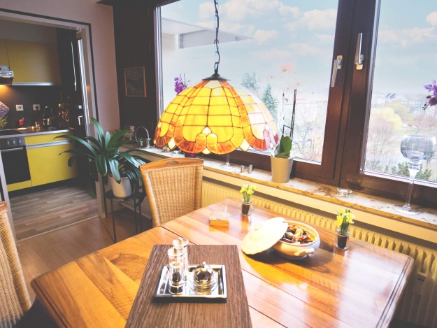 Ein gemütlich eingerichtetes Esszimmer mit einem Holztisch und einer leuchtend orangefarbenen Hängelampe, im Hintergrund ist eine kleine Küche zu sehen.