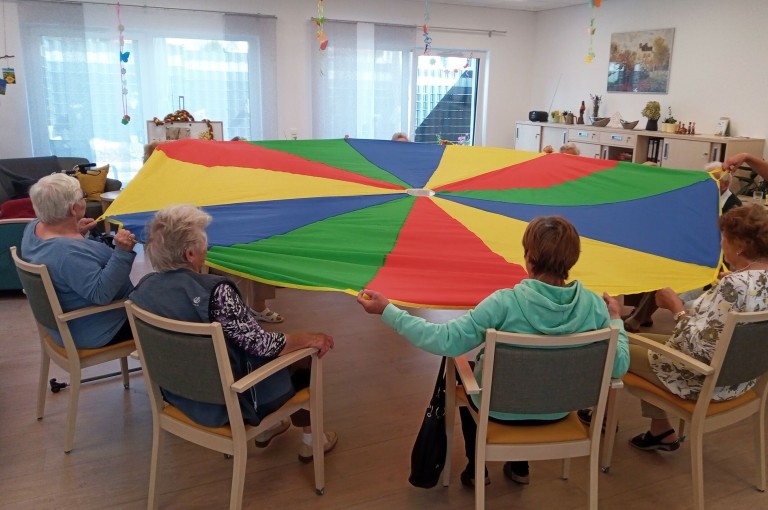Eine Gruppe älterer Menschen sitzt im Kreis und hält gemeinsam ein buntes Schwungtuch gespannt.