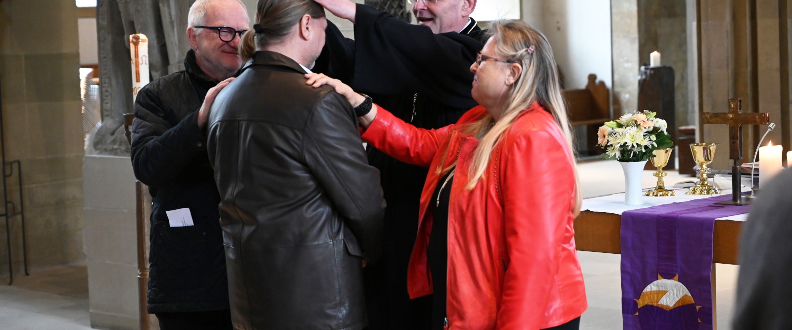 Ein Pfarrer segnet einen Mann in einer Kirche, während zwei weitere Personen ihm die Hände auflegen.