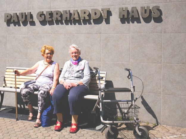 Zwei ältere Damen sitzen lachend auf einer Bank vor der Wand des Paul-Gerhardt-Hauses, auf der der Schriftzug „PAUL GERHARDT HAUS“ angebracht ist. Neben ihnen steht ein Rollator.