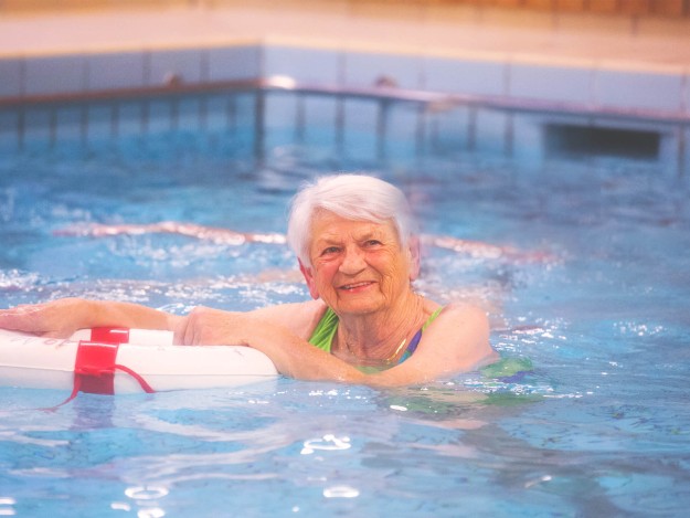 Eine ältere Dame im Wasser, die sich mit einem Schwimmring im Schwimmbad aufhält und fröhlich lächelt.