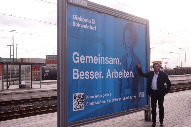 Carsten Bräuer steht vor Plakate mit der Ausschrift Gemeinsam besser Arbeiten