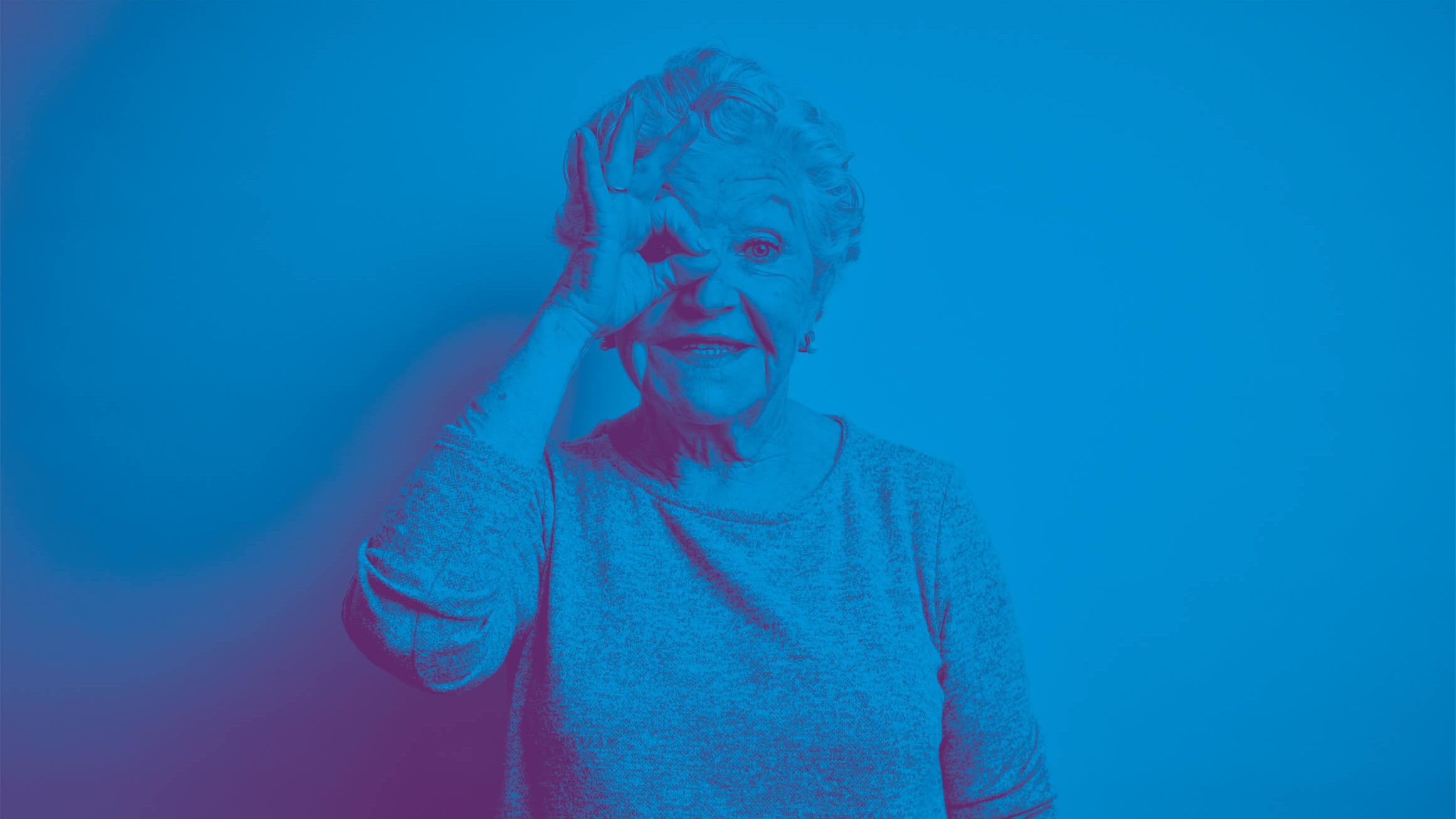 Eine ältere Frau lächelt und formt mit ihrer Hand eine Brille um ihr Auge, als würde sie durch ein Fernglas schauen.