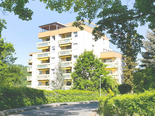 Mehrstöckiges Gebäude mit Balkonen, umgeben von Bäumen und Büschen.