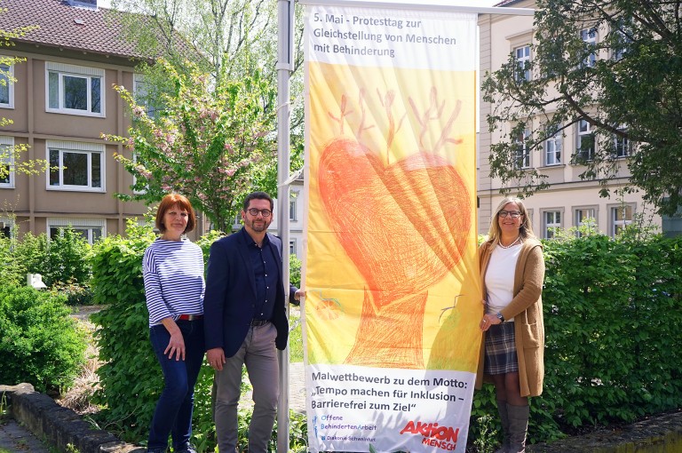 Drei Personen posieren vor einem Banner zum Protesttag für Gleichstellung von Menschen mit Behinderung.