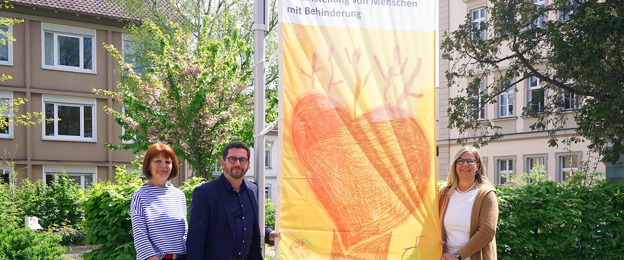 Drei Personen posieren vor einem Banner zum Protesttag für Gleichstellung von Menschen mit Behinderung.