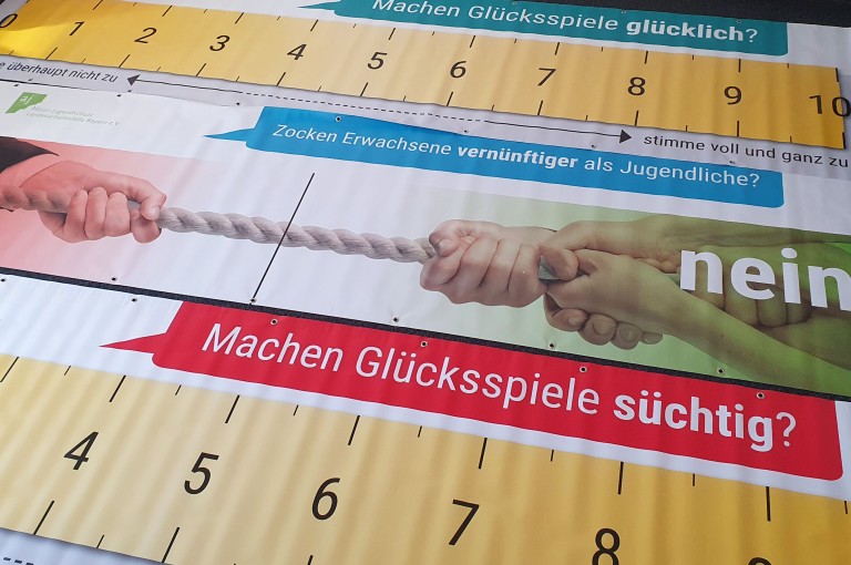 Plakat mit Skalen und Fragen zu Glücksspielsucht, begleitet von einem Bild zweier Hände im Tauziehen.