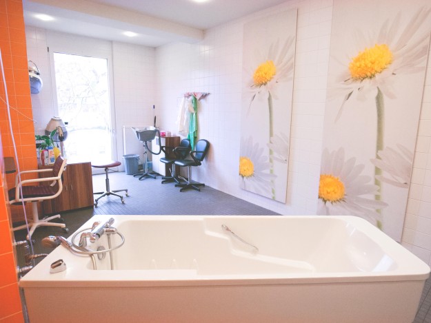 Heller Raum mit Badewanne, Friseurstühlen und großen Blumenbildern an den Wänden.