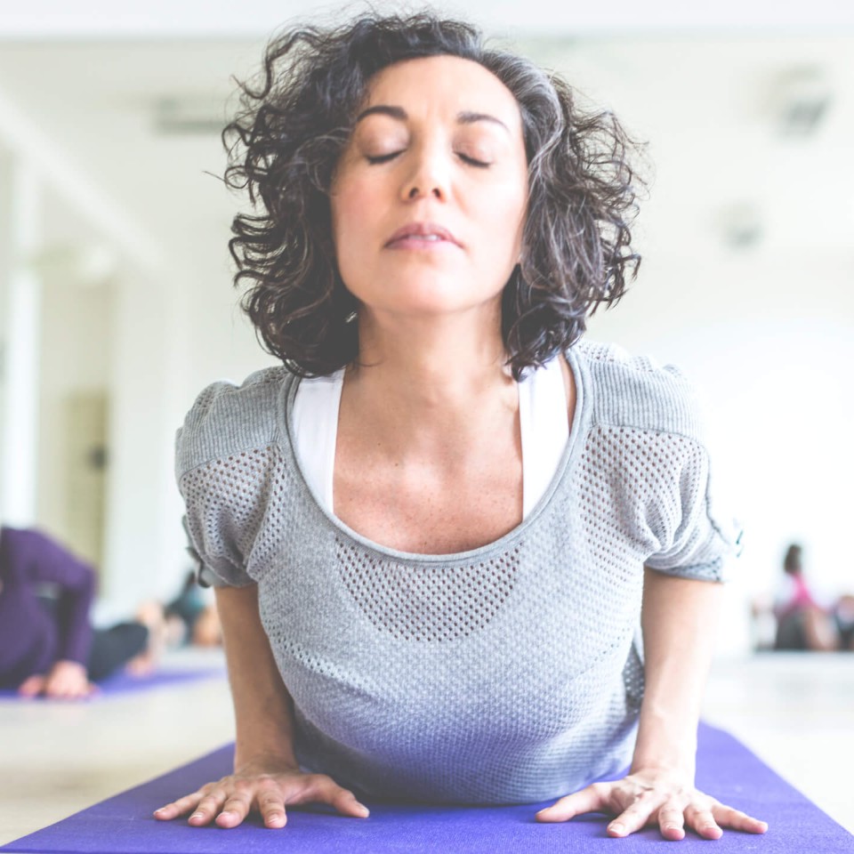 Eine Frau macht eine Yogaübung auf einer Matte, die Augen geschlossen, im Hintergrund weitere Teilnehmer.